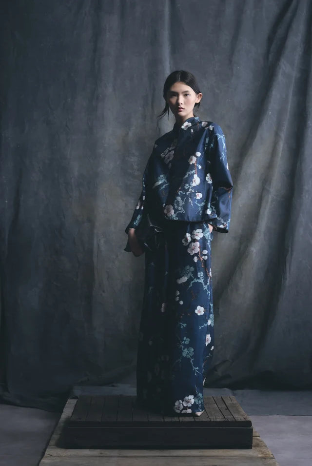 A model stands on a wooden platform wearing a dark navy blue kimono-style outfit with a white and pink floral print, set against a dark gray fabric backdrop.