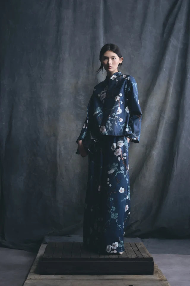 A model stands on a wooden platform wearing a dark navy blue kimono-style outfit with a white and pink floral print, set against a dark gray fabric backdrop.